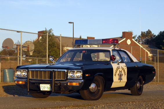 Black and white 1973 Dodge 1500 police car with light bar, parked on gravel lot