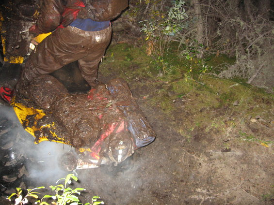 Mud-covered yellow and red ATV partially visible, with a person in muddy brown gear standing over it