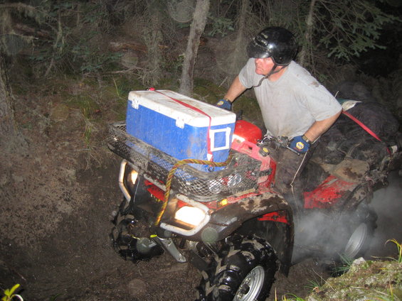 Red ATV with a blue and white cooler strapped to the rear rack, navigating a muddy trail