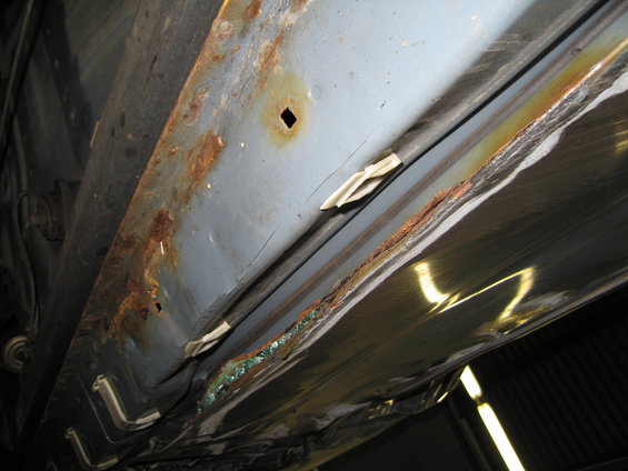 Close-up of heavily rusted metal on a vehicle's undercarriage, showing corrosion and damage along the rocker panel