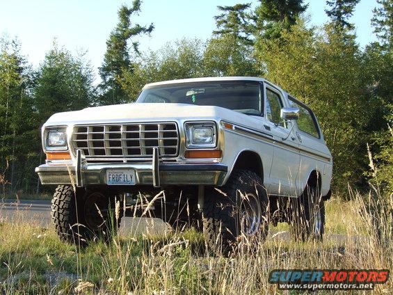 White 1978 Ford Bronco with lifted suspension, off-road tires, and custom license plate 'FRDFILY', parked in grassy area