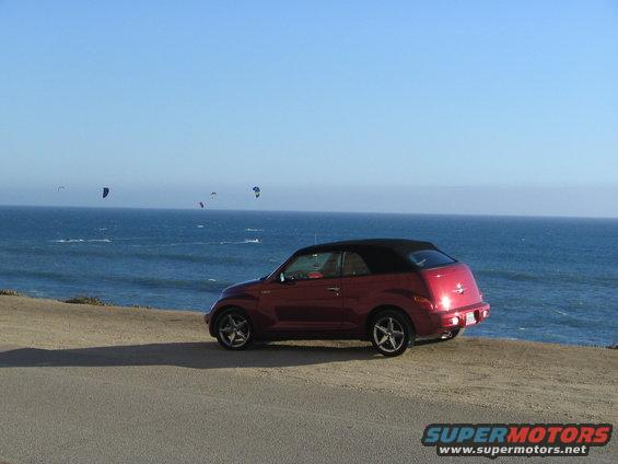 Red convertible PT Cruiser parked on a dirt road overlooking the ocean, with kitesurfers in the distance