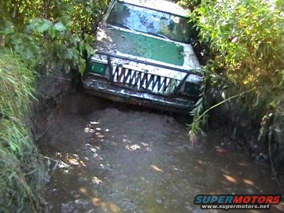 Mud-covered off-road vehicle with a shark-tooth grille stuck in a muddy ditch