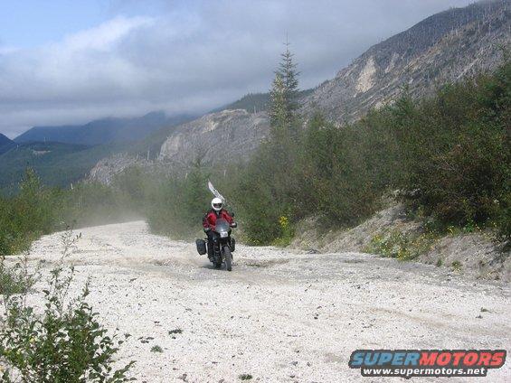 Motorcyclist riding on a dusty, gravel road with mountains in the background