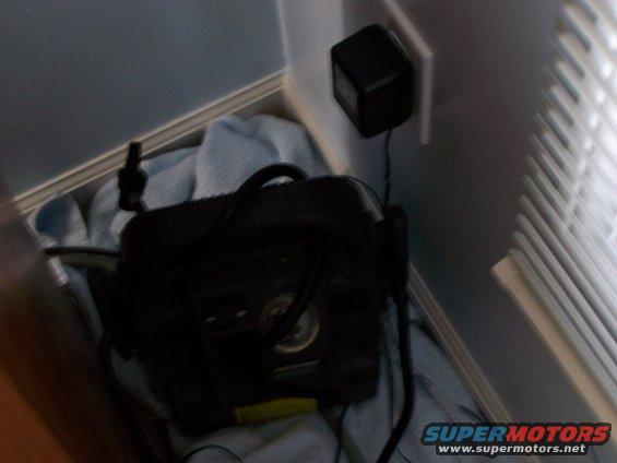 Black air compressor with gauge and hoses, plugged into a power adapter on a wall