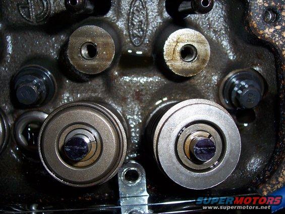 Close-up of engine cylinder head with valves and springs visible. Some bolts and metal parts are also in view