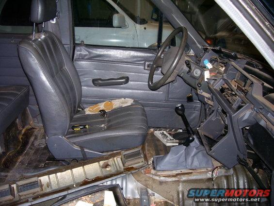 Interior of a vehicle with seats and dashboard removed, showing exposed metal and wiring. Another white vehicle is visible in the background
