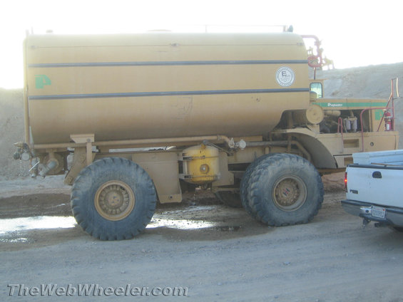 Big yellow water truck with huge tires parked next to a white pickup truck