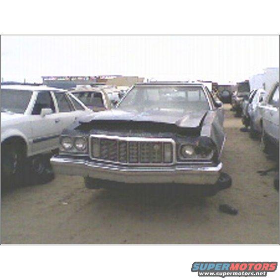 Front view of a dark 1973 Ford Ranchero with a damaged headlight and grille, parked in a junkyard next to other vehicles