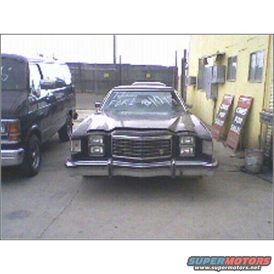 Front view of a dark sedan parked next to a van. Text on windshield reads 'FAKE 81095'. Signs in background say 'CARS FOR SALE'