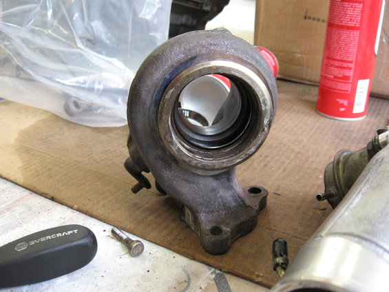 Turbocharger housing with visible internal fins and mounting flange. A red spray can and a screwdriver are nearby
