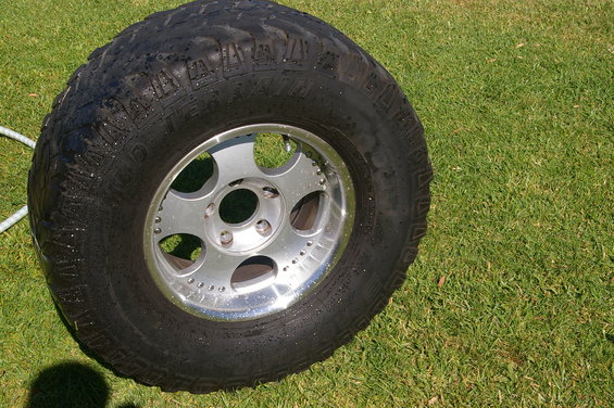 Close-up of a large off-road tire on a silver rim, sitting on green grass. Some water droplets are visible on the tire