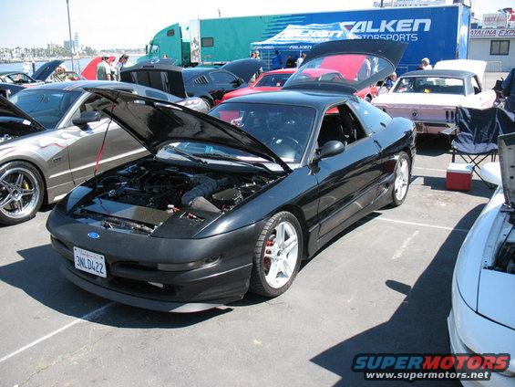 Black 1995 Ford Probe with hood open, parked at an outdoor car show. Other cars and a Falken Motorsports truck are visible