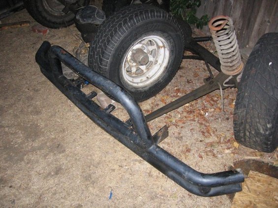 Black off-road bumper and spare tire with white rim, resting on the ground near a wooden fence