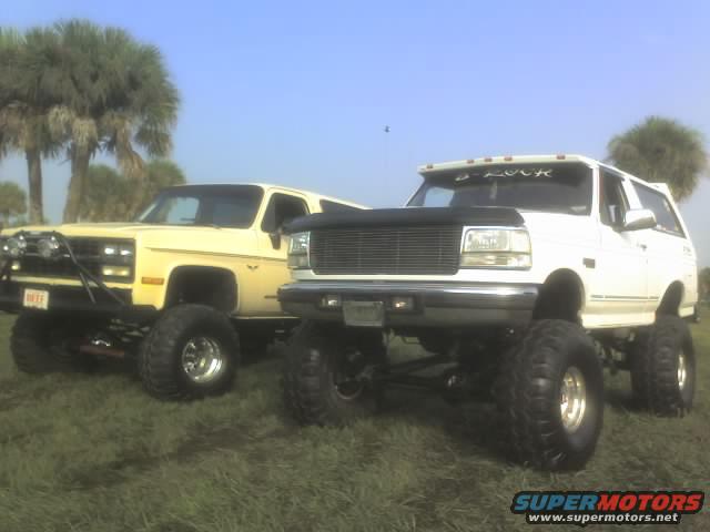 White Ford Bronco and yellow Chevy Blazer, both lifted with large tires, parked on grass
