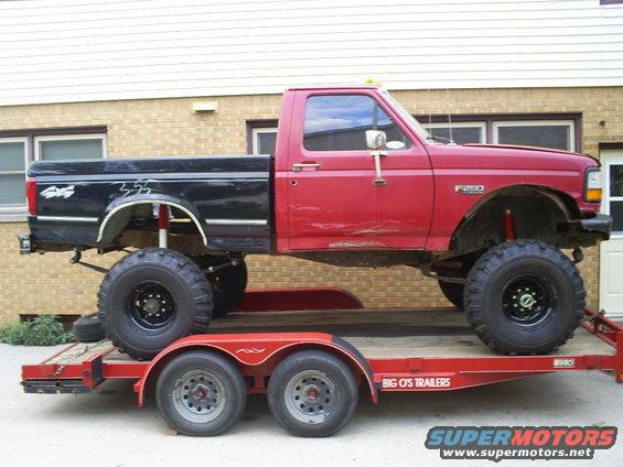 1994 Ford F-250 pickup truck, lifted with large tires, red cab and black bed, on a red trailer