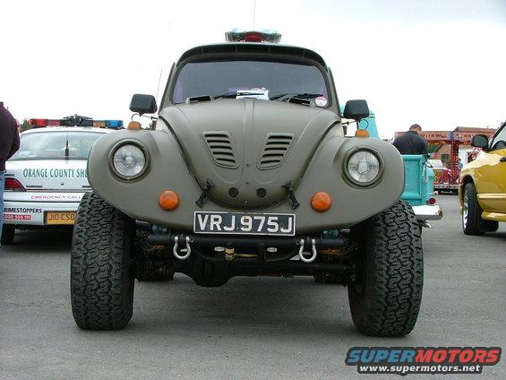 Front view of a lifted, olive-drab Volkswagen Beetle with off-road tires. A police car and a yellow truck are in the background