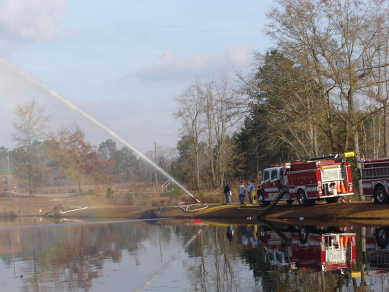 Two red fire trucks are parked on the bank of a lake, with one spraying a powerful stream of water into the lake. Several people stand near the trucks
