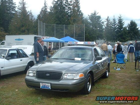 2000 Ford Crown Victoria police car with light bar, parked on grass next to a white Mustang
