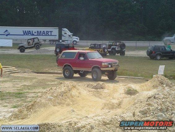 Red Ford Bronco with lifted suspension on dirt track, other Jeeps and Walmart truck in background