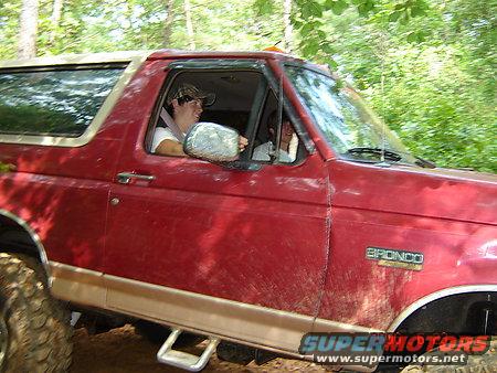 Red Ford Bronco with a person driving, off-roading in a wooded area. The Bronco badge is visible