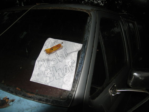 Windshield of a vehicle with a sign that reads 'The Miobi Shop' and a yellow glove attached