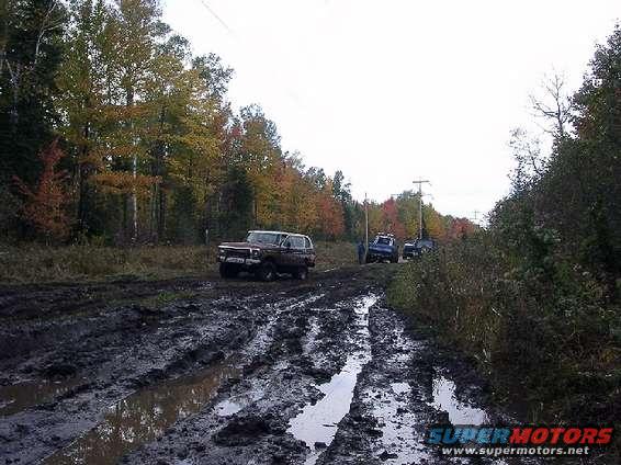 brown SUV and two other vehicles on muddy track with trees in background