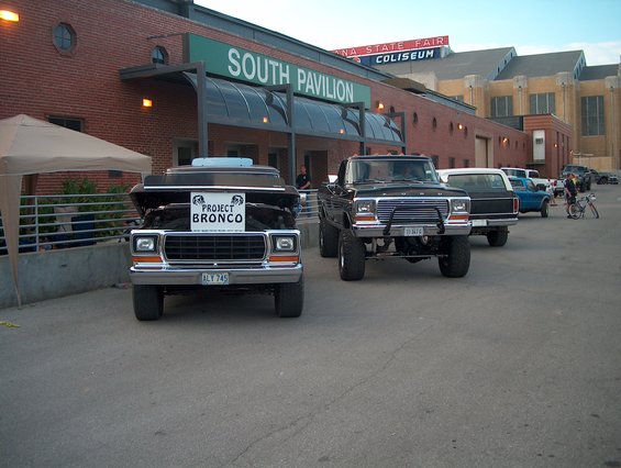 Front view of a black 1978 Ford Bronco with hood up and "PROJECT BRONCO" sign, parked next to a lifted black Ford pickup truck