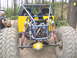 1989 Ford Bronco - A custom buggy with a yellow body and blue roll cage sits on a dirt path surrounded by trees.