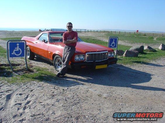 Orange coupe utility parked by a beach with a man leaning on it. Two handicap parking signs are visible