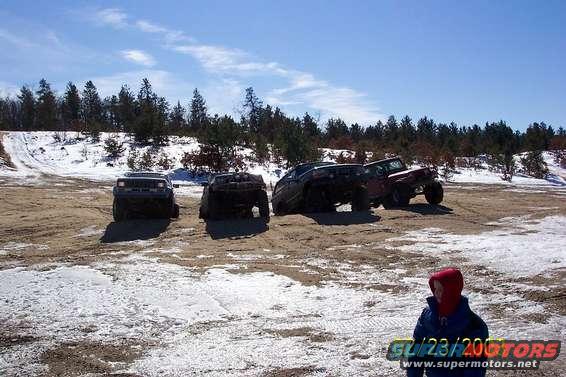 Four-wheel drive vehicles on a dirt and snow-covered hill under a blue sky