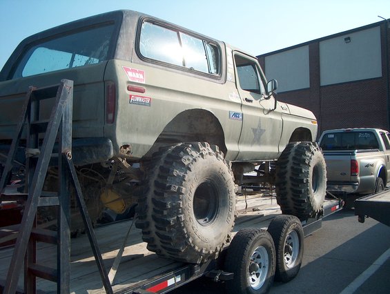 Olive green 1978 Ford Bronco with large off-road tires on a trailer, next to a pickup truck