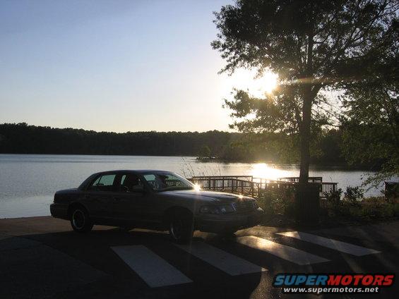 Dark sedan parked near a lake at sunset, with a tree and wooden dock in the background