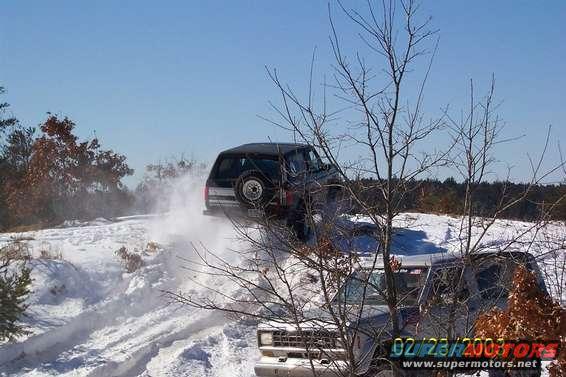 Two SUVs driving through snow. One is kicking up snow as it goes up a hill