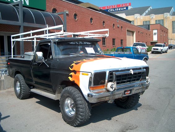 Black and white pickup truck with flames on hood, ladder rack, and large tires. Blue and white trucks in background