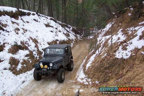 Black Jeep CJ-7 driving on a muddy, snow-dusted dirt road through a wooded area