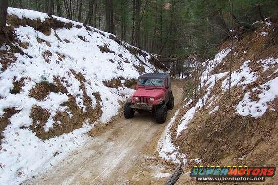 Red Jeep Wrangler with large tires on a muddy, snow-dusted trail