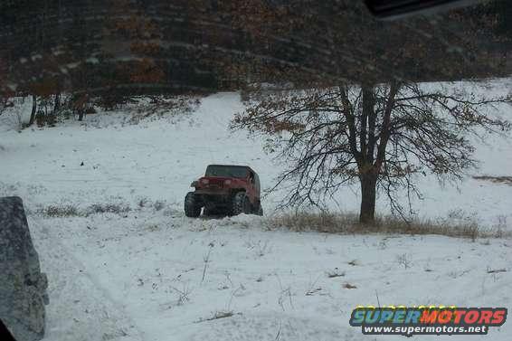 Red Jeep Wrangler driving in a snowy field next to a bare tree