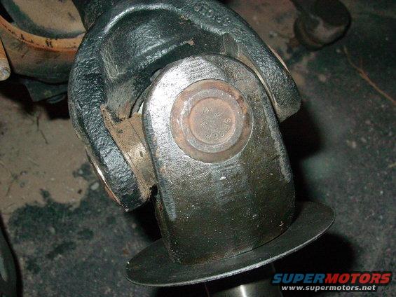 Driveshaft U-joint with visible grease and rust, showing a circular metal cap with stamped text