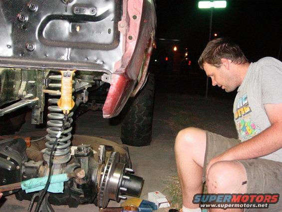Man working on the front suspension of a large vehicle, possibly a truck or SUV. The vehicle's fender and frame are visible