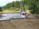 1989 Ford Bronco - A dark SUV navigates a water-filled dirt track surrounded by trees.