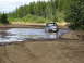 1989 Ford Bronco - A dark-colored 1980s Ford F-Series truck navigates through a muddy water obstacle in a wooded area.