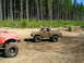 1989 Ford Bronco - Two off-road trucks navigate a muddy path through a wooded area.