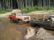 1989 Ford Bronco - Two off-road trucks are stuck in a muddy water crossing in a wooded area.