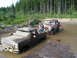 1989 Ford Bronco - Two pickup trucks are stuck in a muddy area with trees in the background.