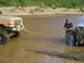 1989 Ford Bronco - Two trucks stuck in a muddy water hole, one being pulled out with a yellow strap.