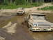 1989 Ford Bronco - Two off-road vehicles, a pickup truck and an SUV, navigate a muddy water obstacle on a wooded trail.