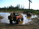 1989 Ford Bronco - Two trucks are stuck in a muddy water pit surrounded by trees and clear sky.