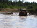 1989 Ford Bronco - A red off-road vehicle navigates through a muddy, water-filled terrain with dense trees lining the horizon.