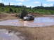 1989 Ford Bronco - A dark pickup truck navigates a muddy water obstacle on a sunny day, surrounded by trees and dirt mounds.
